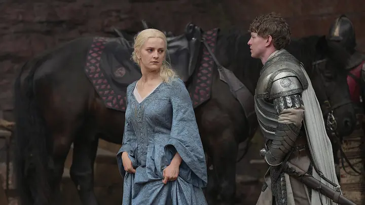 Phia Saban as Helaena Targaryen pulling up her blue gown while talking to a Kingsguard knight beside horses in House of the Dragon.