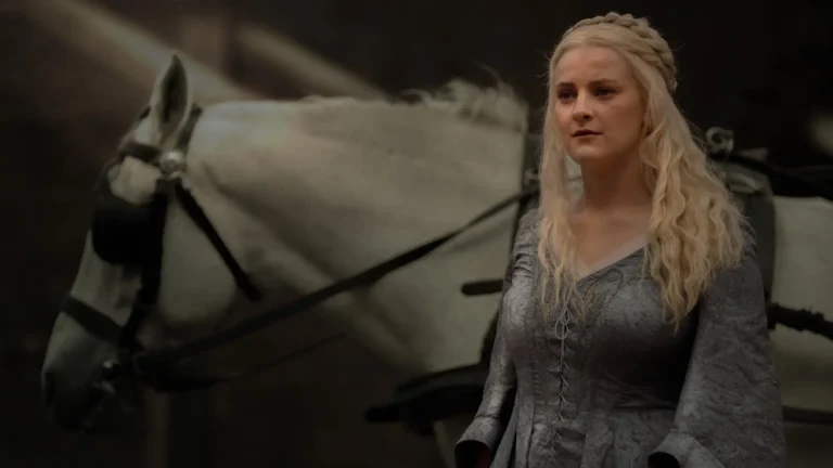 Phia Saban as Helaena Targaryen wearing a grey gown standing beside a white horse in House of the Dragon.