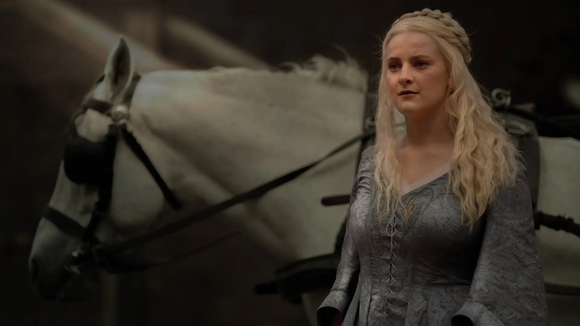 Phia Saban as Helaena Targaryen wearing a grey gown standing beside a white horse in House of the Dragon.