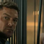 Jack Reacher, a muscular man with short brown hair and a furrowed brow, stares intensely forward through vertical metal bars.