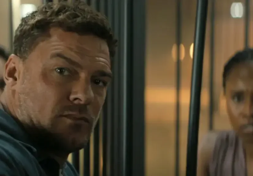 Jack Reacher, a muscular man with short brown hair and a furrowed brow, stares intensely forward through vertical metal bars.