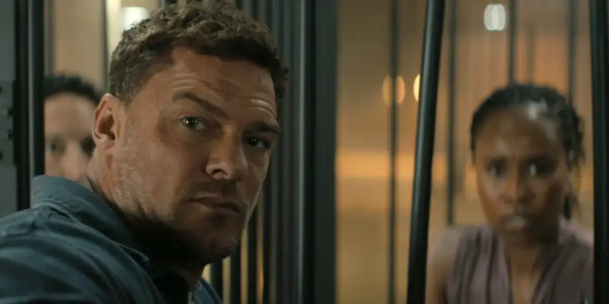 Jack Reacher, a muscular man with short brown hair and a furrowed brow, stares intensely forward through vertical metal bars.