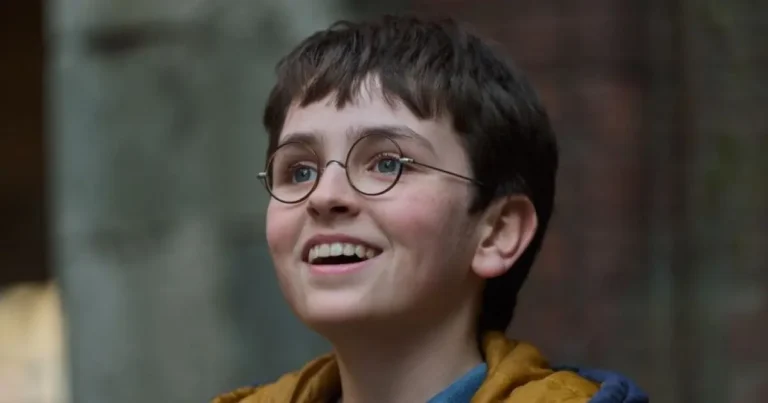 A young boy with short brown hair and round wire-rimmed glasses smiling upward cheerfully.
