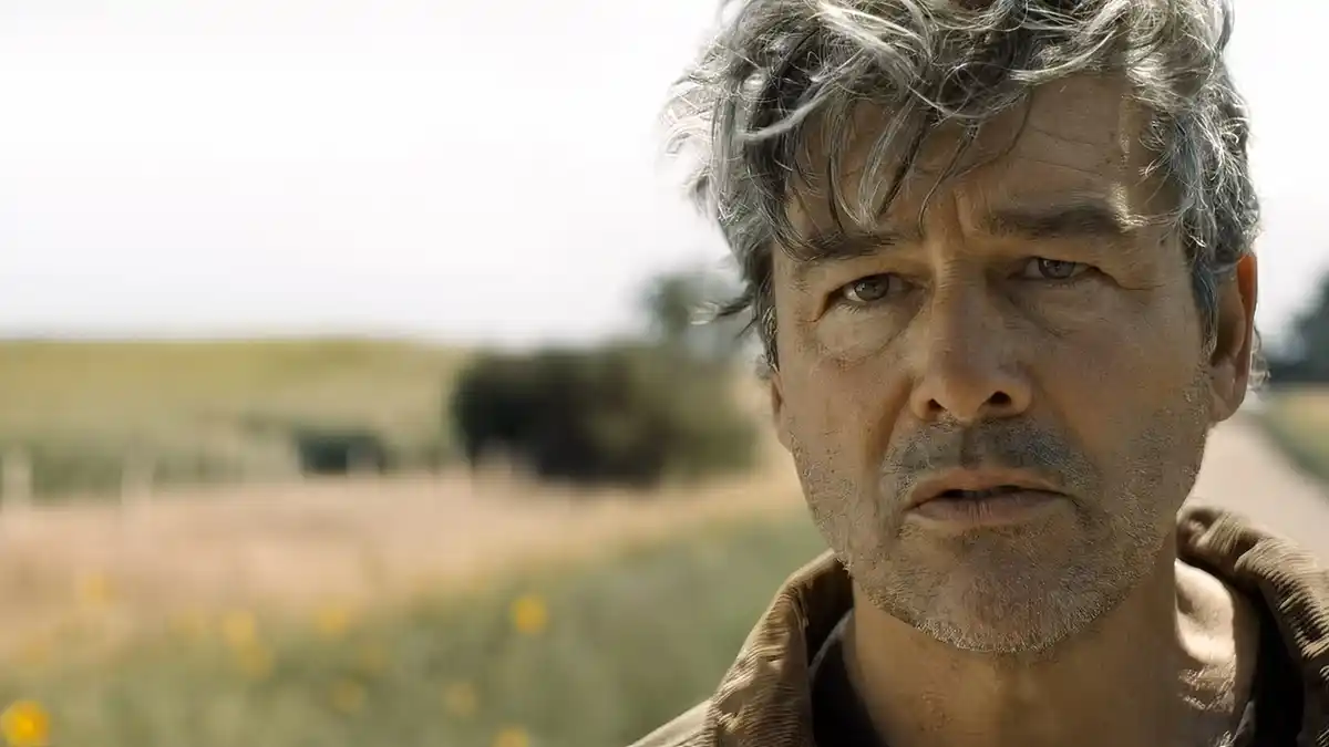 Close-up of a middle-aged man with messy gray hair, slight stubble, and a somber expression, standing outdoors against a blurred rural landscape.