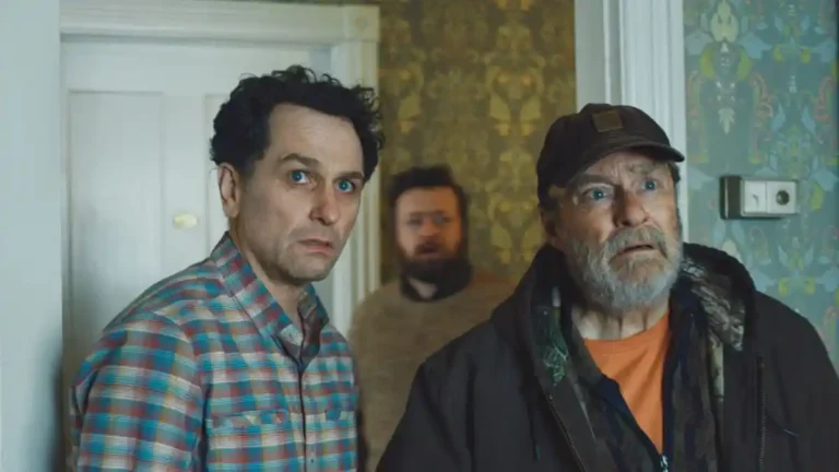Three men stand inside a room with patterned wallpaper, staring off-camera with wide eyes and expressions of shock and disbelief.